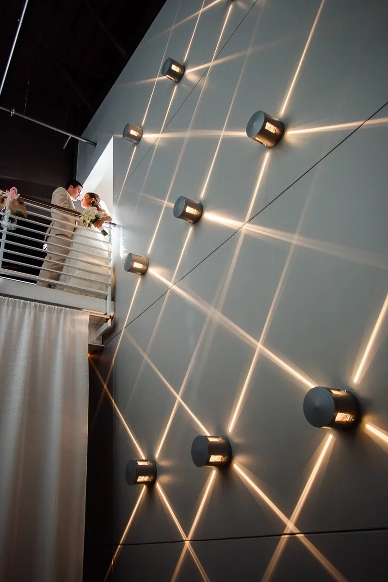 A couple stands on a balcony, looking down with joy. The wall behind them features modern lighting with crisscrossing beams, creating a sophisticated ambiance.