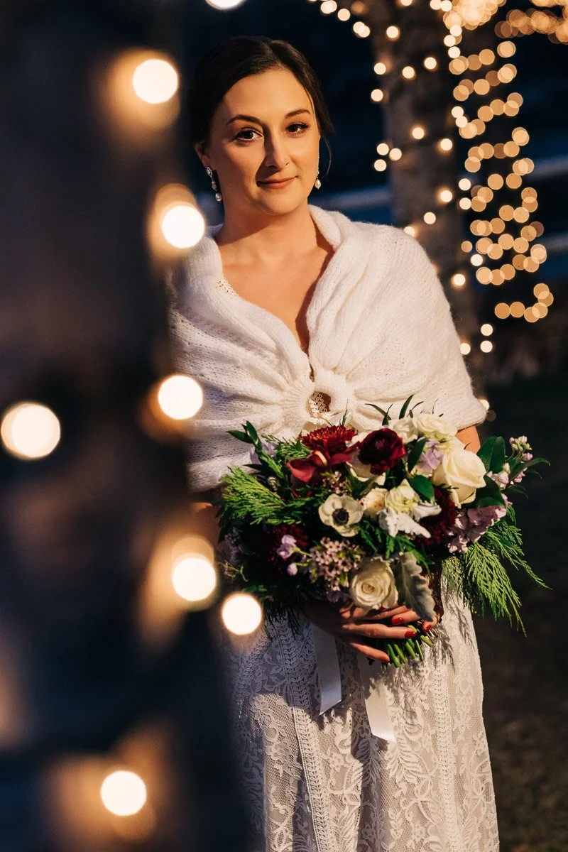 Elegant bride in a white lace dress and shawl holds a vibrant bouquet. Soft light from tree fairy lights creates a warm, romantic ambiance.
