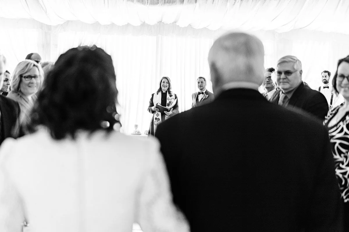 A wedding ceremony in black and white. A couple walks down the aisle towards smiling guests and an officiant, creating a joyful and celebratory atmosphere.