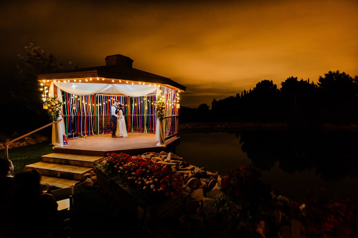 A couple is dancing on a decorated gazebo at night, illuminated by warm string lights. The sky is a deep orange, reflecting over a calm pond. Romantic atmosphere.