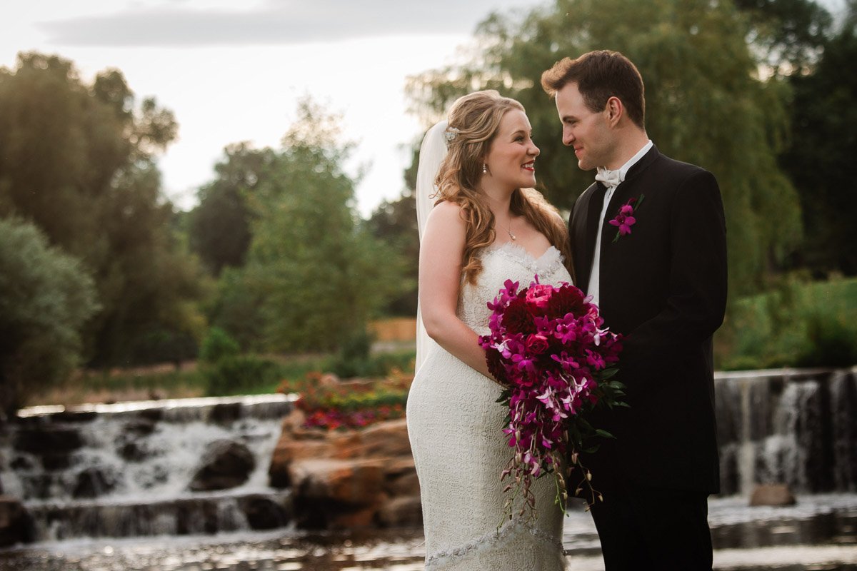 A bride in a white lace gown and veil holds a vibrant bouquet, standing with her groom in a black suit by a serene waterfall, sharing joyful smiles.