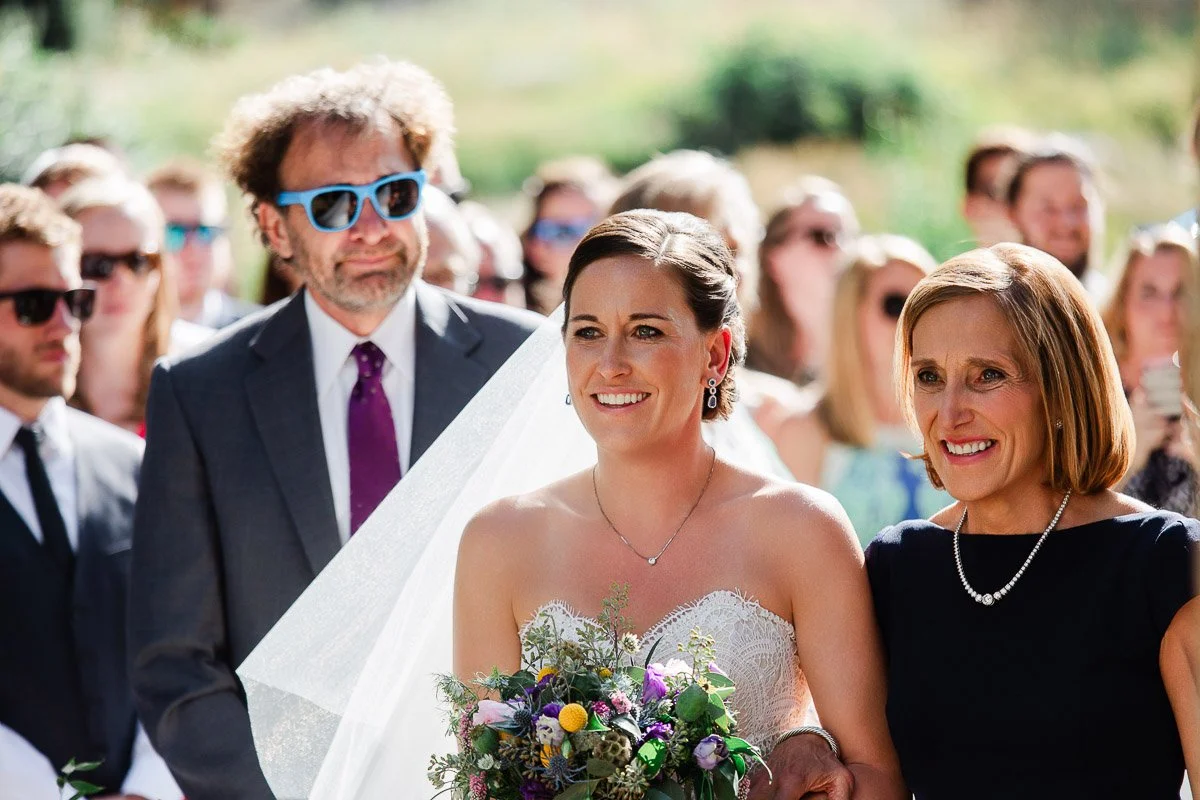 Bride in white dress, veil, and holding a bouquet stands smiling between two people in a sunlit outdoor ceremony, surrounded by guests. Joyful atmosphere.