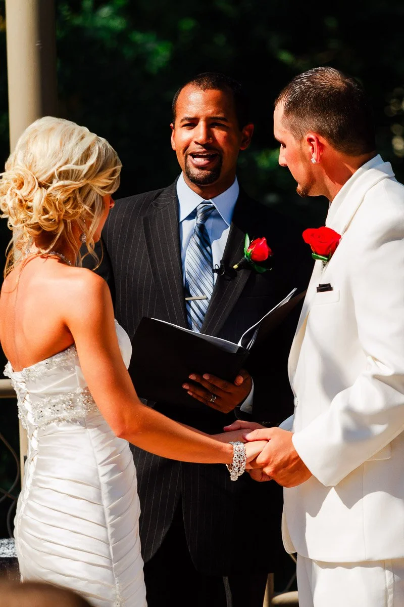 A couple in formal attire exchanges vows, holding hands at an outdoor wedding. The officiant, in a suit, smiles warmly. The scene feels joyful and intimate.