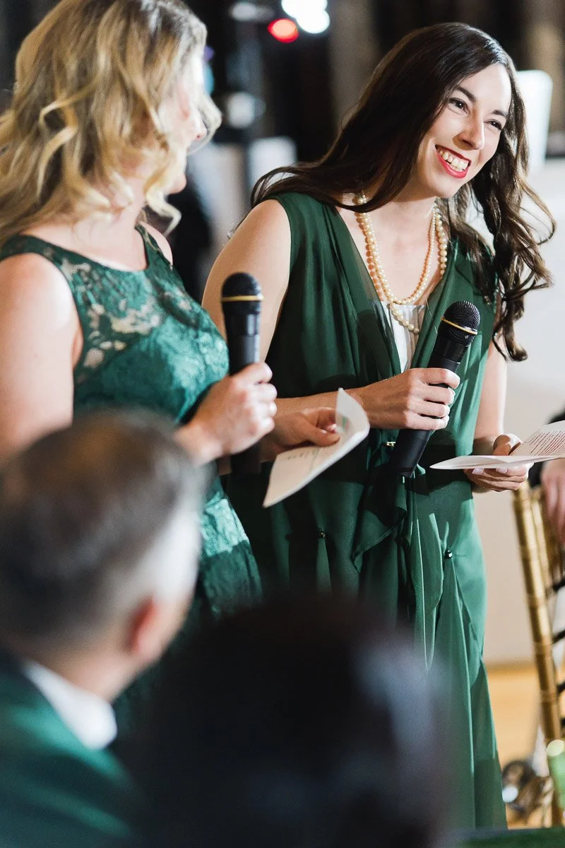 Two women in green dresses hold microphones and papers, smiling warmly while speaking at an event. People are seated in the foreground, listening attentively.