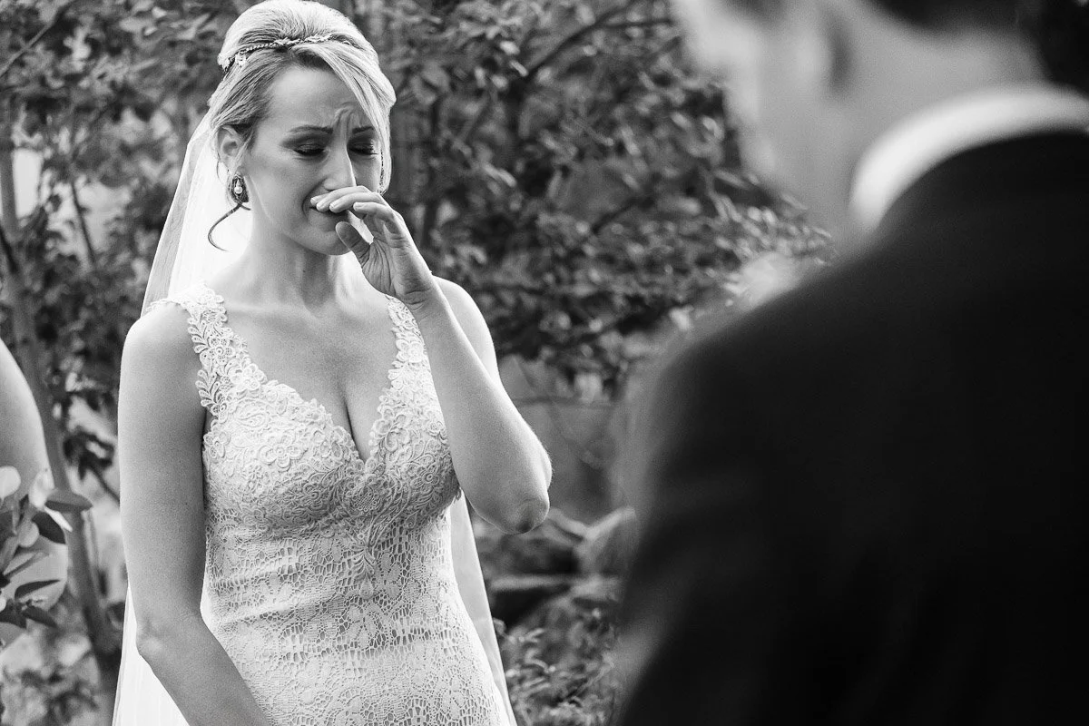 A bride in a lace gown wipes tears during an emotional outdoor ceremony. Her expression is touching, surrounded by greenery, with the groom blurred nearby.