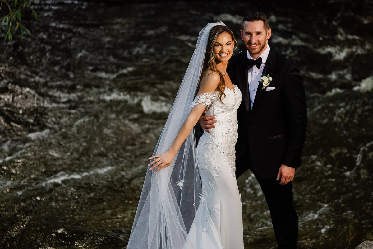 A bride and groom stand in front of a flowing river. The bride wears a white gown and veil, while the groom is in a black tuxedo. Both are smiling joyfully.