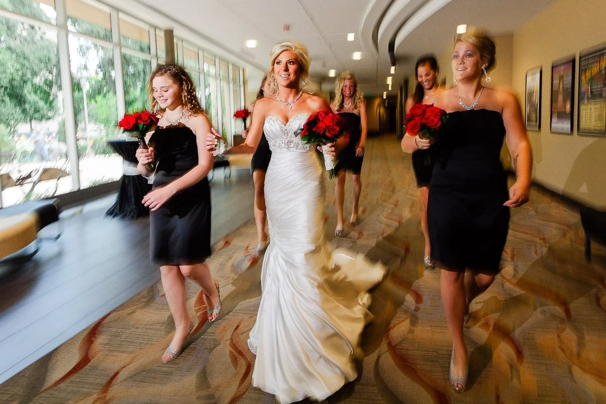 A bride in a flowing white gown walks with bridesmaids in black dresses during a Lincoln Center wedding holding red bouquets down a lit hallway, exuding joy and anticipation.