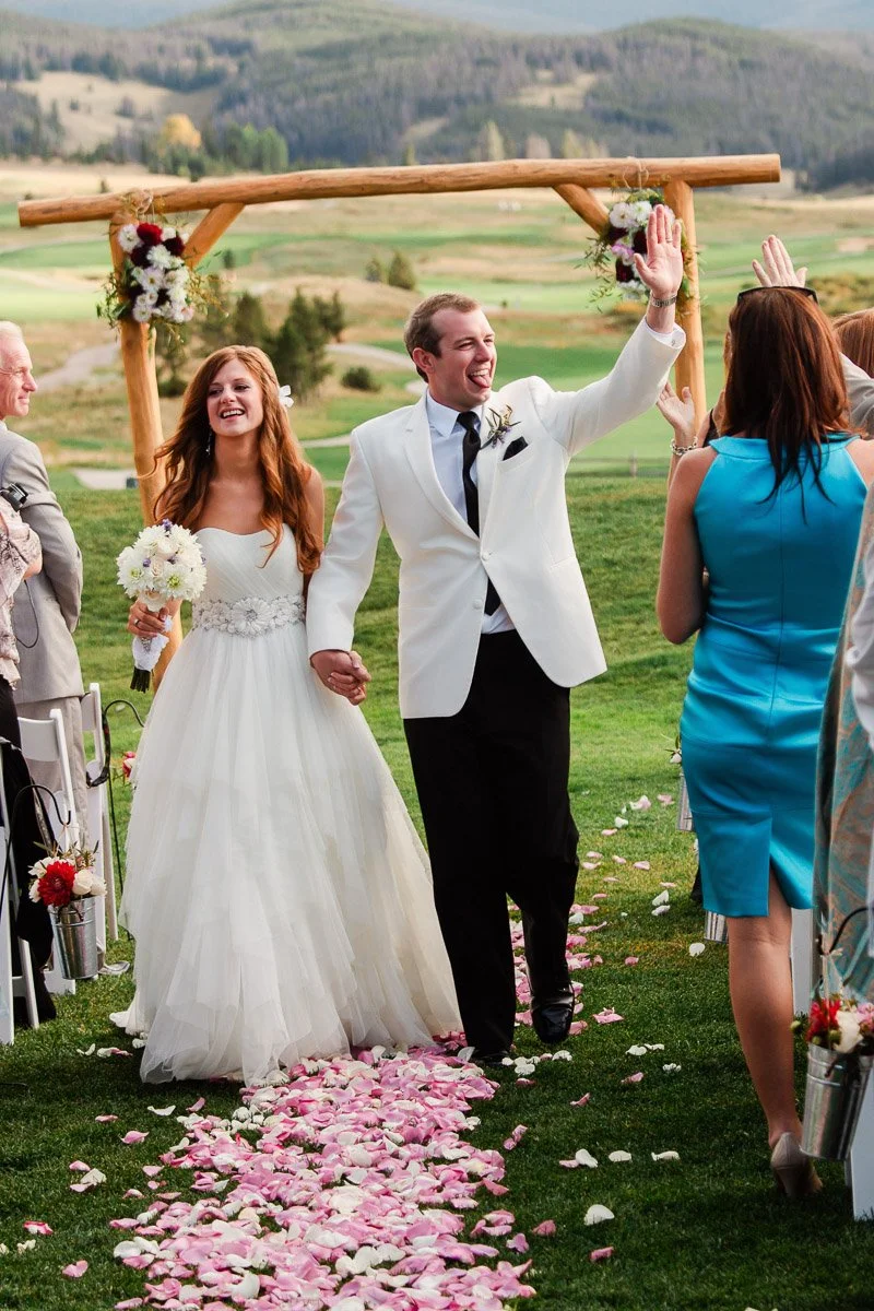 Bride in a white gown and groom in a white jacket joyfully walk down a petal-strewn aisle, under a floral arch, in a scenic outdoor setting captured by keystone wedding photographer tomKphoto