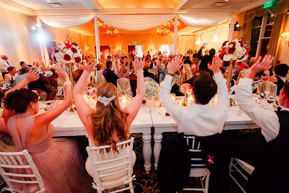 Guests at a formal event raise their hands in celebration, seated at a white-decorated table adorned with flowers, under soft, warm lighting.
