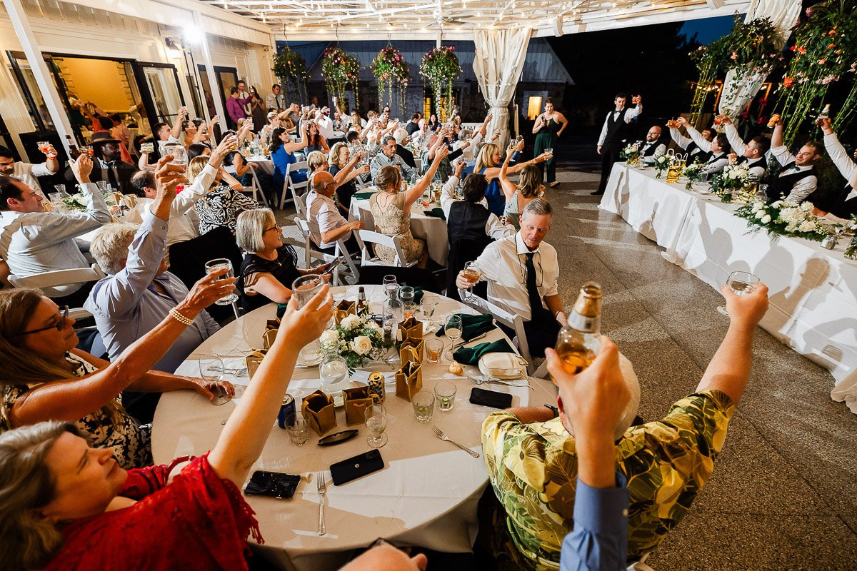 A joyful wedding reception with guests raising glasses in a toast. Tables are adorned with flowers and the room is warmly lit, creating a festive atmosphere.