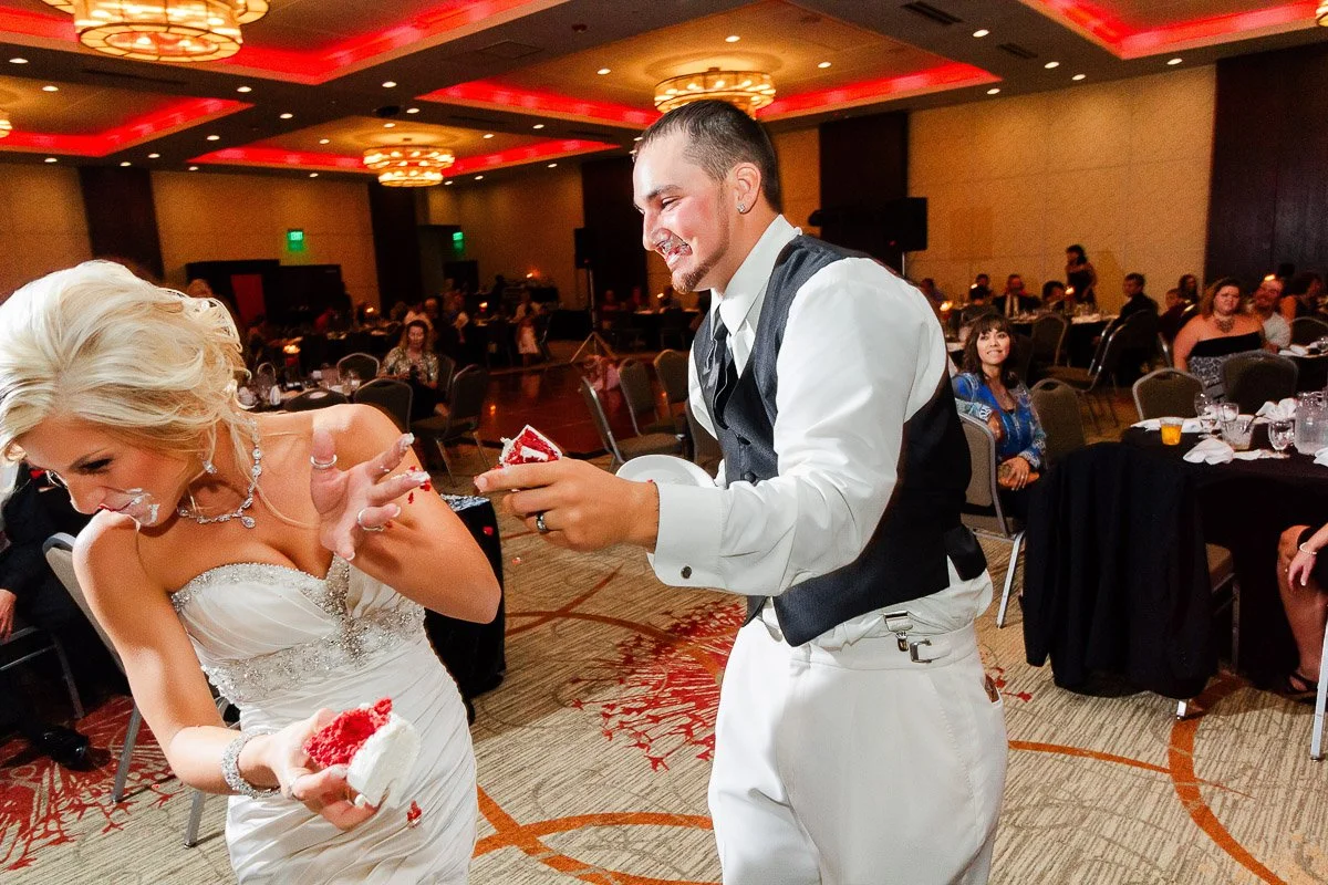 A bride and groom in a humorous cake-smashing moment. Both have cake on their faces, laughing joyfully. Guests and a warmly lit reception hall are in the background.