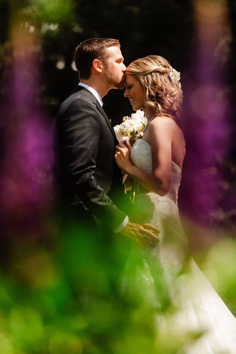 Bride and groom share a tender moment outdoors. The groom kisses the bride's forehead, framed artistically by blurred purple and green foliage during a Donovan Pavilion wedding in Vail, Colorado