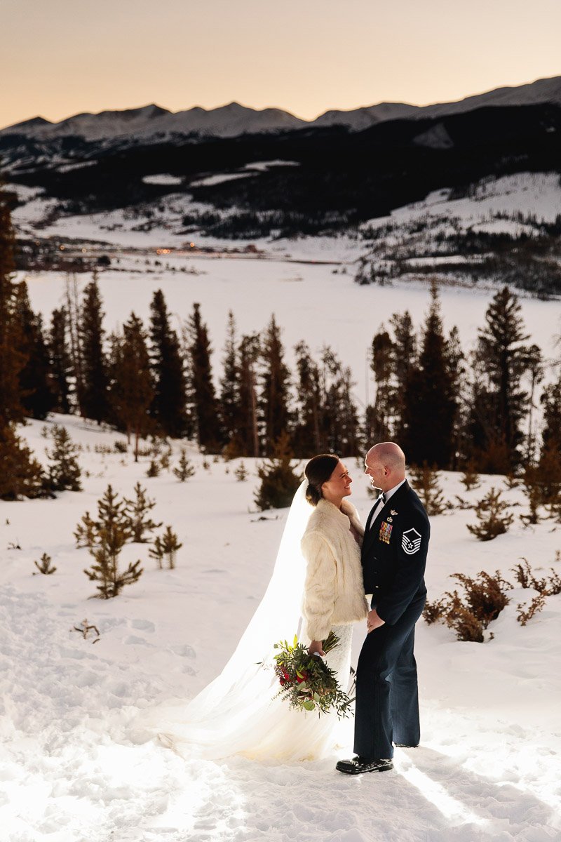A bride in a fur jacket and a groom in a military uniform stand smiling in a snowy landscape with mountains at sunset. The atmosphere is romantic and joyful.