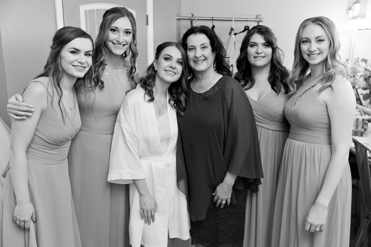 A joyful black-and-white photo of six women dressed in bridesmaid gowns and a bride in a robe. They stand closely together, smiling warmly at the camera.
