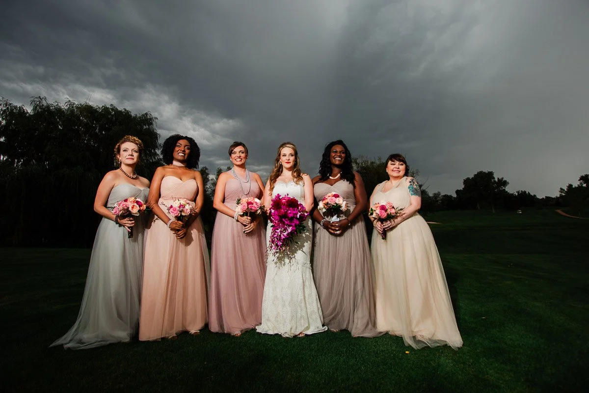 A bride in a white dress holds a bouquet of pink flowers, standing with five bridesmaids in pastel gowns under a dramatically cloudy sky.