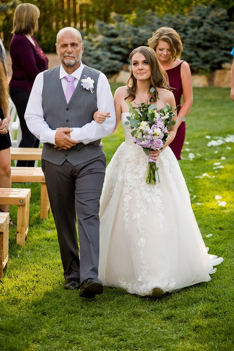 A bride in a lace gown holds a bouquet while being escorted down a grassy aisle by a man in a vest and tie. Guests and trees are visible in the background.
