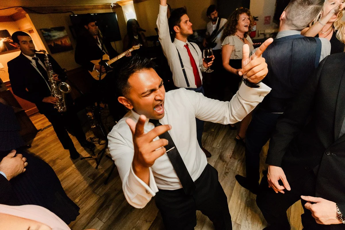A lively party scene with a man in a tie enthusiastically dancing in the foreground, surrounded by smiling guests and a live band playing in the background.