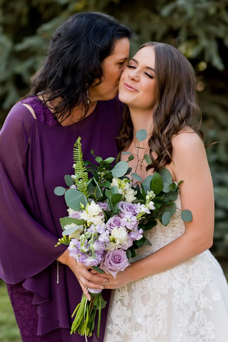 A woman in a purple dress kisses a smiling bride holding a bouquet of purple and white flowers. The scene conveys warmth and joy outdoors.
