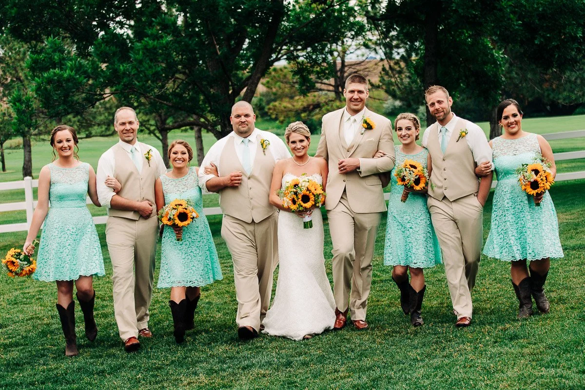 A joyful wedding party poses outdoors on grass, with the bride in a white gown flanked by bridesmaids in turquoise dresses and groomsmen in beige suits, all holding sunflowers.