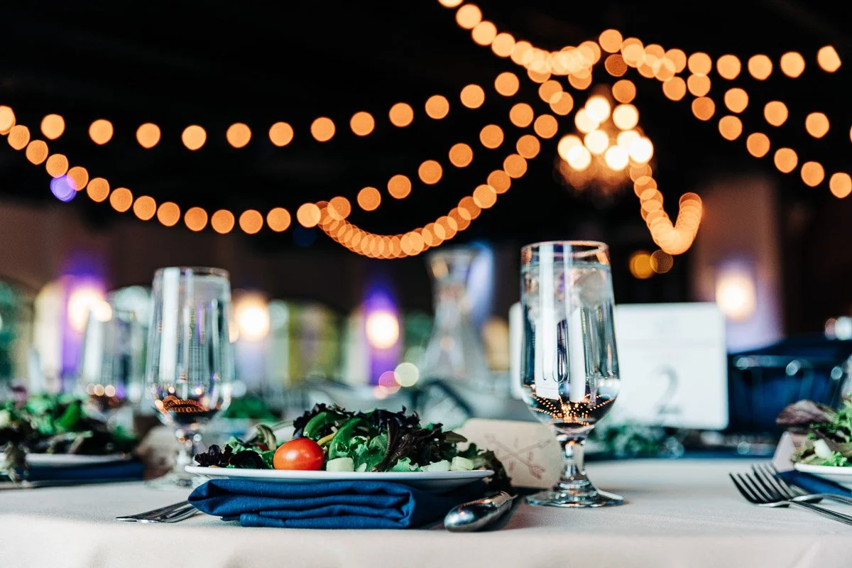Elegant table setting with a salad and glasses on a white tablecloth, under warm string lights creating a cozy, festive atmosphere.