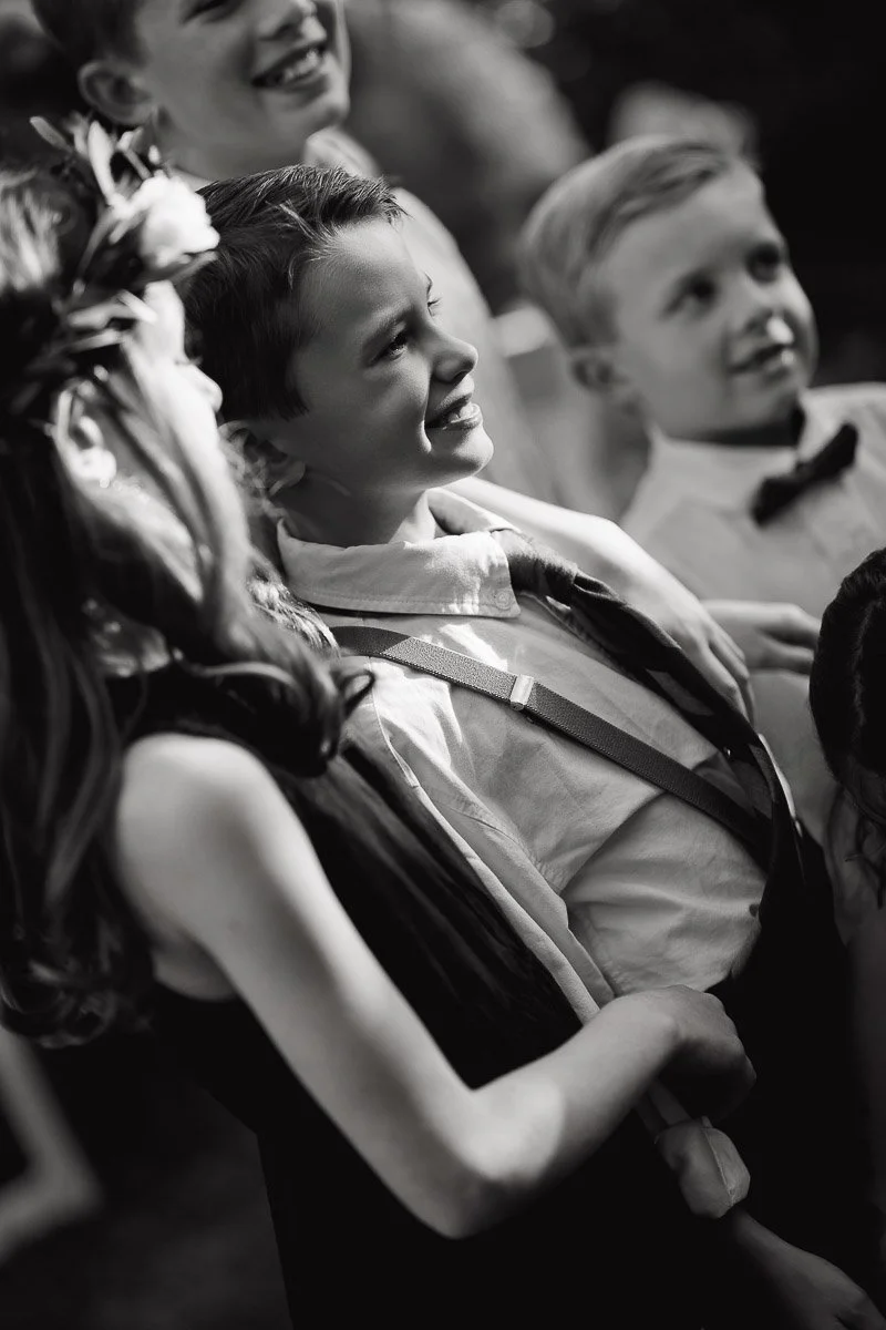 Black and white photo of smiling children dressed in formal attire, capturing joy and innocence. A girl with flowers in her hair stands closest.