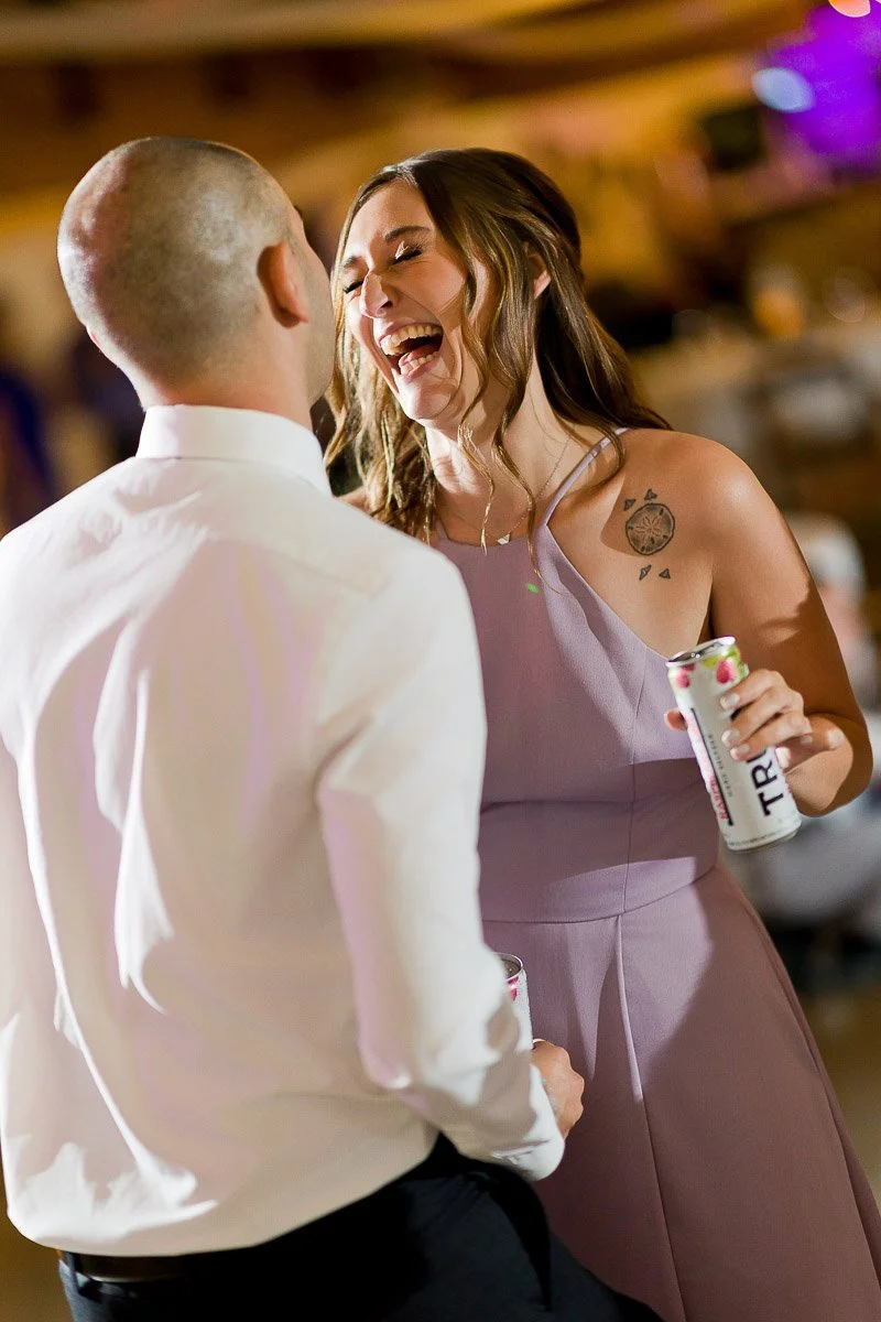 A woman in a lavender dress laughs joyfully with a man in a white shirt at a lively event, both holding drinks, conveying happiness and celebration.