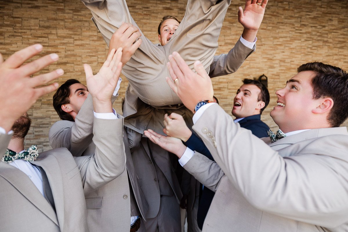 A group of joyful men in suits toss another man into the air against a brick wall backdrop. The atmosphere is celebratory and lively.