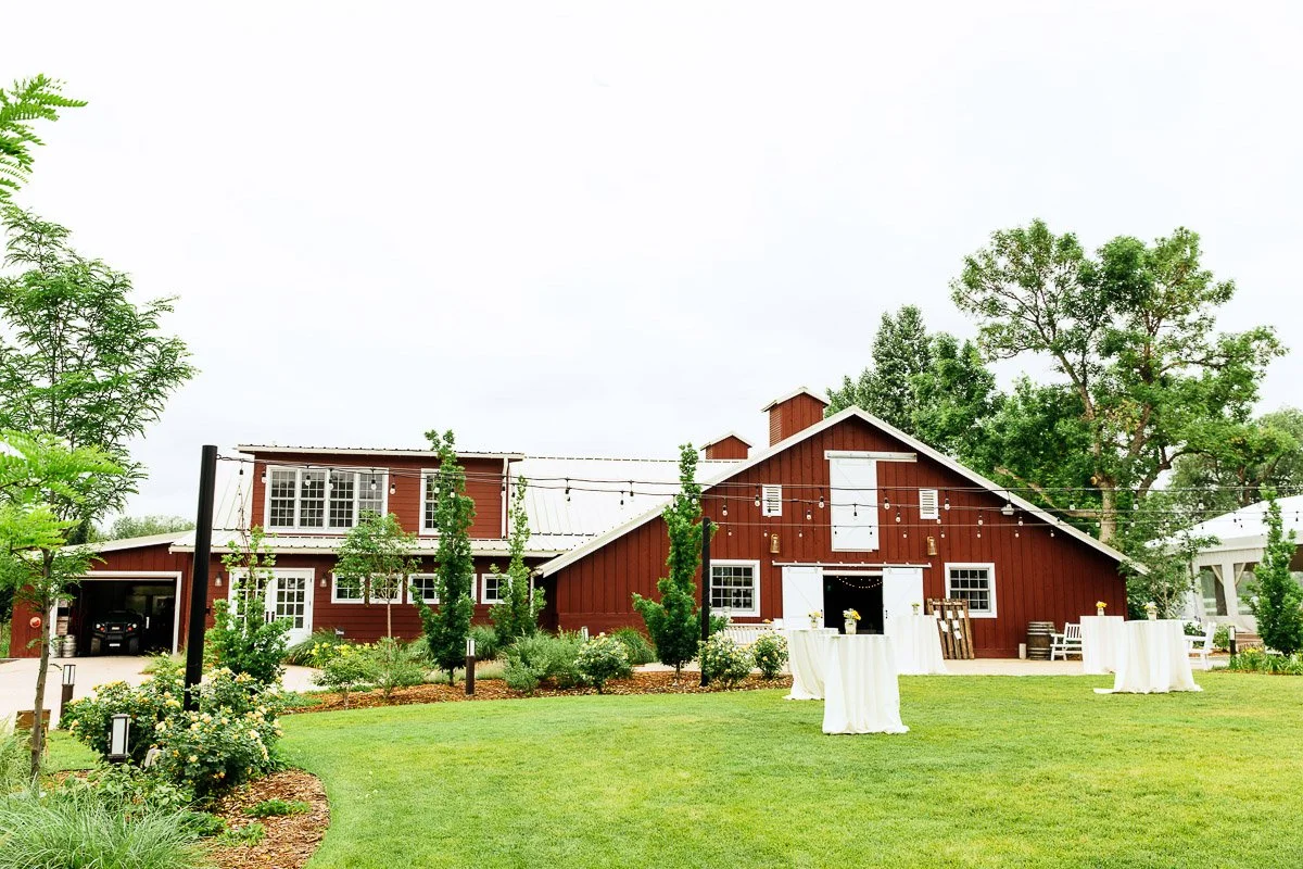 Red barn venue with string lights and white tables on lush grass, surrounded by trees and greenery, conveying a rustic and inviting atmosphere.
