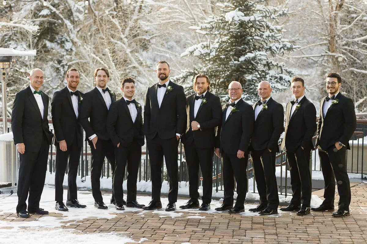 A group of ten men in black tuxedos and bow ties stand smiling in a snow-covered outdoor setting. The mood is formal and celebratory.