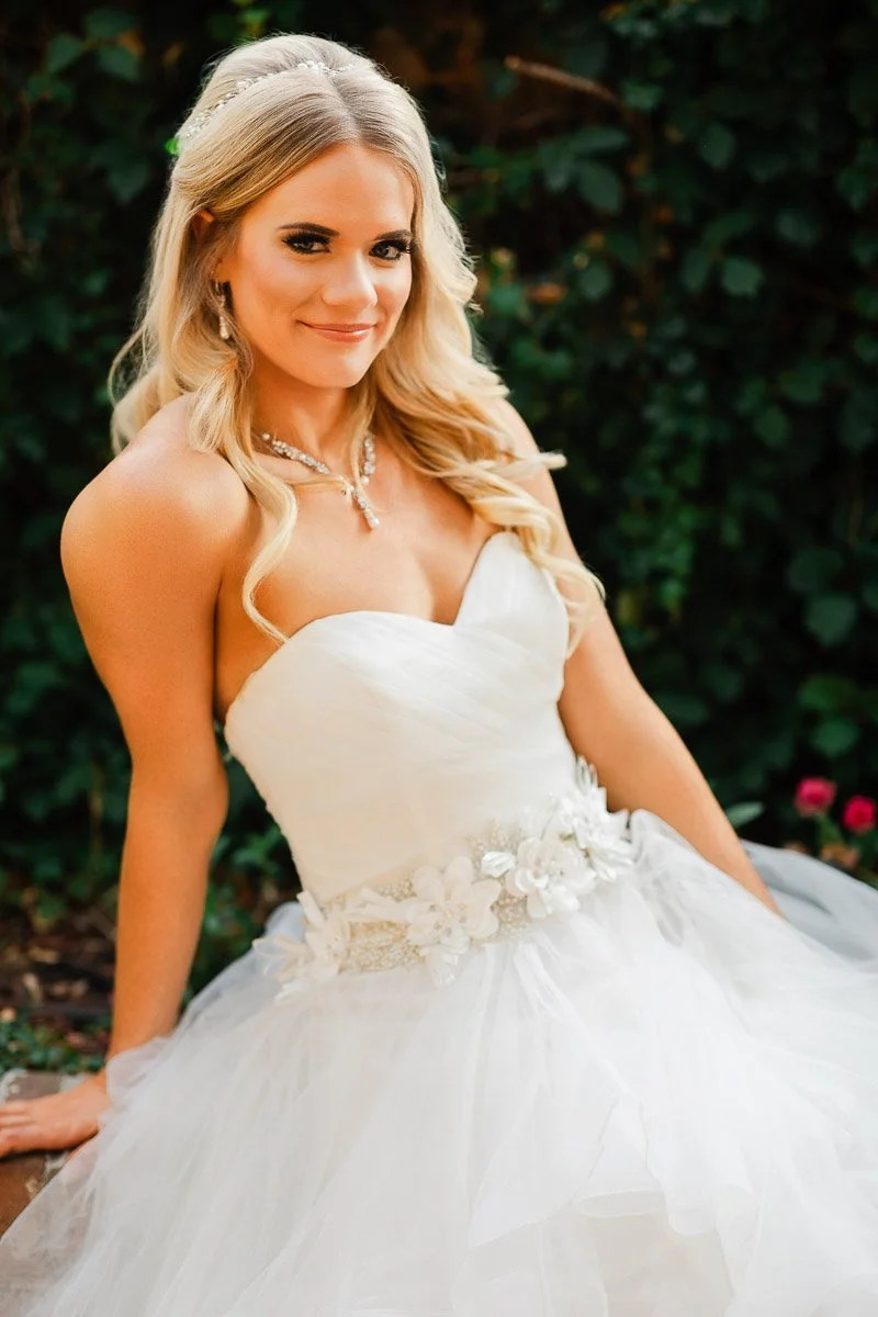 A smiling bride in a strapless white gown with a floral belt sits against a green leafy backdrop. Her long blonde hair is styled in soft waves.