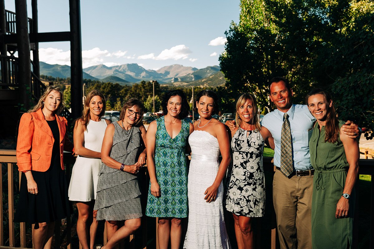 A group of smiling people, dressed in summer attire, stands together on a patio with a mountain range and trees in the background, conveying happiness.