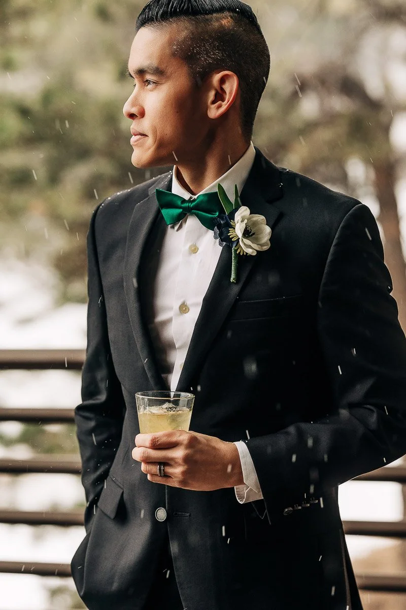 A man in a black suit with a green bow tie and boutonniere holds a drink, gazing thoughtfully to the side. Snow falls gently around him, suggesting a serene winter setting.