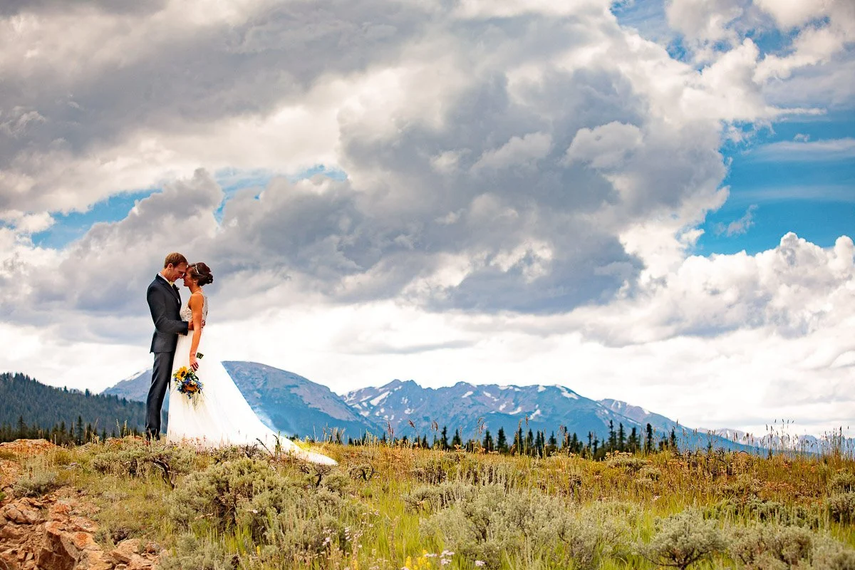 A couple in wedding attire stands on a grassy hill at a Keystone Ranch wedding, embracing under a dramatic sky with scattered clouds. Mountains create a majestic backdrop, conveying romance.