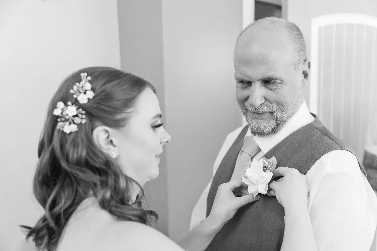 A woman with styled hair pins a flower on a smiling man's vest. They share a tender moment, suggesting a wedding or special occasion. Black and white photo.