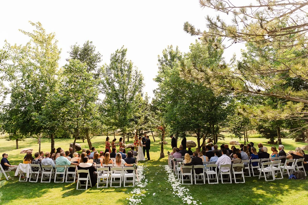 Outdoor wedding ceremony in a lush garden with rows of seated guests. The couple stands under trees, surrounded by greenery and scattered white petals.