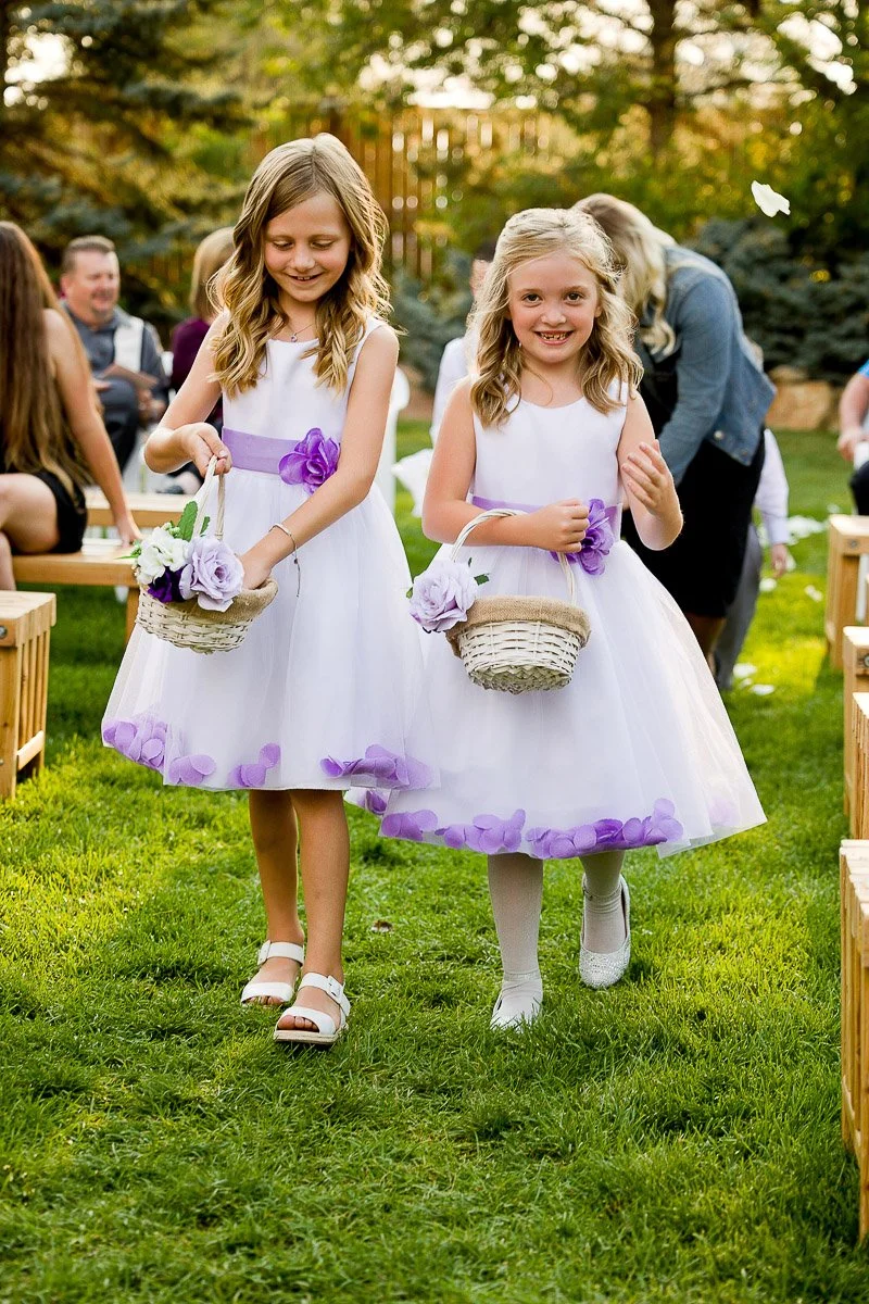 Two young flower girls in white dresses with purple accents walk on grass, holding baskets with flowers. They smile, creating a joyful wedding scene.