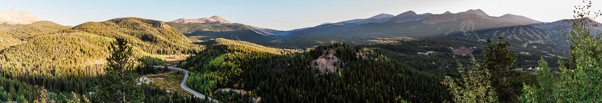 Panoramic view of a mountain landscape with sunlit, tree-covered hills and a winding road. The sky is clear, suggesting a calm and serene atmosphere.