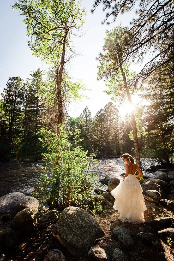 A bride stands by a serene forest river in sunlight, surrounded by rocks and tall trees. The scene evokes tranquility and natural beauty.