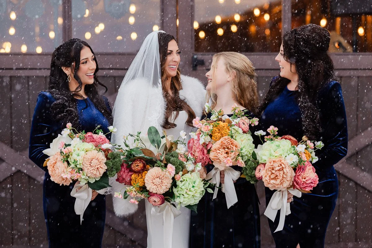 Bride in white and bridesmaids in blue velvet hold colorful bouquets, smiling joyfully in falling snow. Warm lights glow softly in the background.