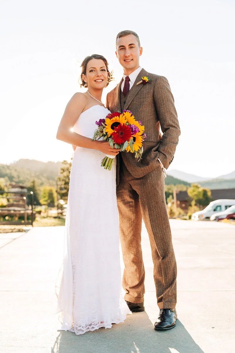 A smiling couple in wedding attire stands outdoors in sunlight. The bride holds a vibrant bouquet of sunflowers and daisies, with a scenic mountain backdrop.