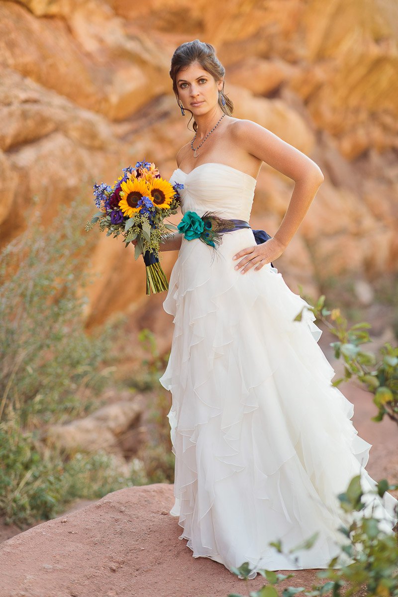 Bride in a flowing white dress poses against rocky backdrop, holding vibrant sunflower bouquet. Her expression is serene and confident.