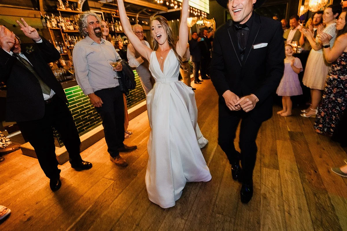 A joyful bride in a white dress dances with a groom in a black tuxedo at a lively reception. Guests cheer in a warmly lit, festive atmosphere.