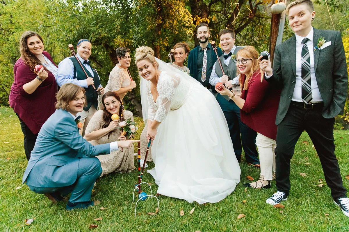 A joyful bride in a white gown plays croquet on grass, surrounded by smiling friends holding mallets. The scene is lively and celebratory.
