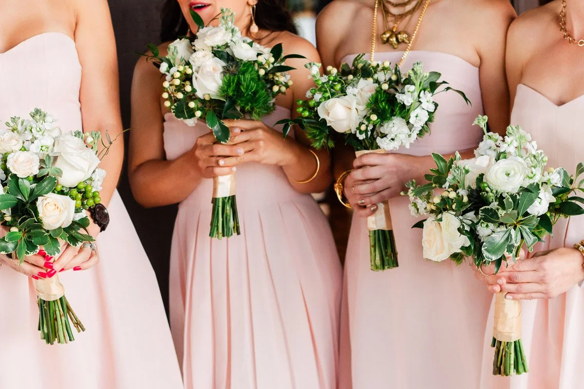 Four bridesmaids in soft pink dresses hold matching bouquets of white and green flowers, conveying elegance and joy.