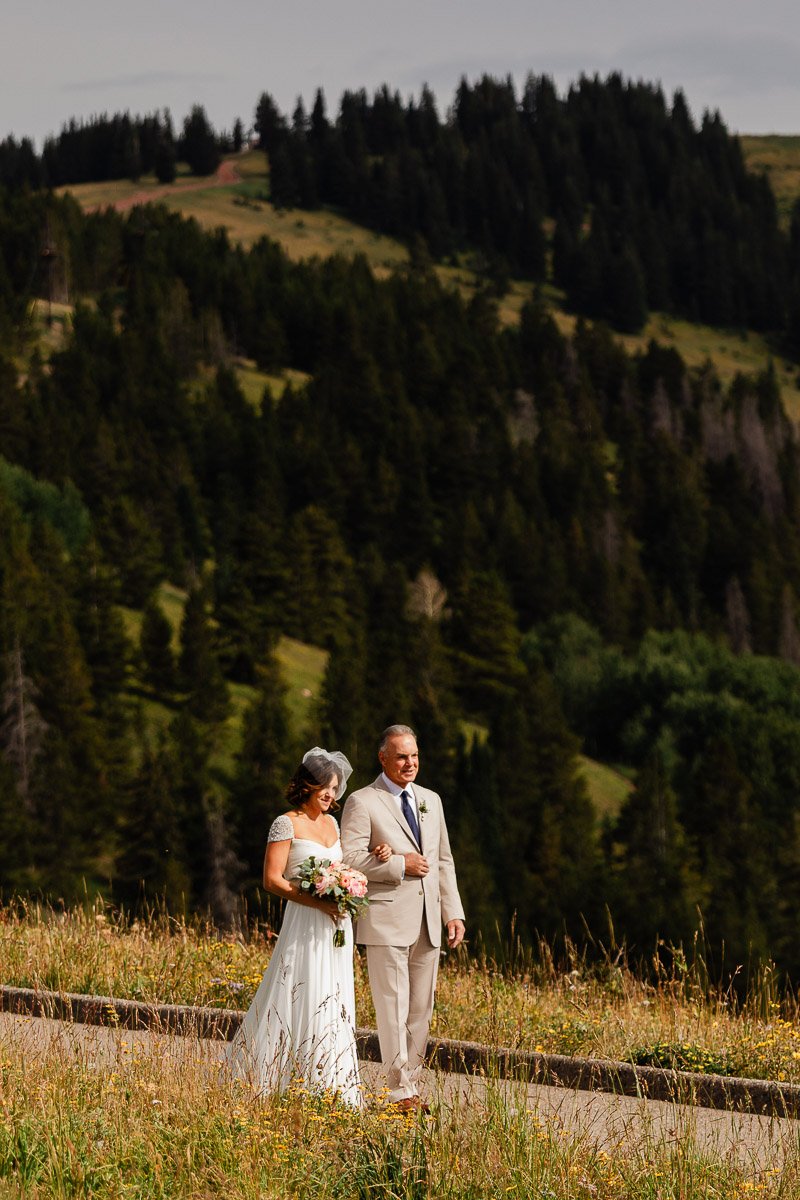 A bride in a white dress walks with an older man in a light suit down a grassy path, surrounded by lush green hills and trees, conveying joy and tranquility.