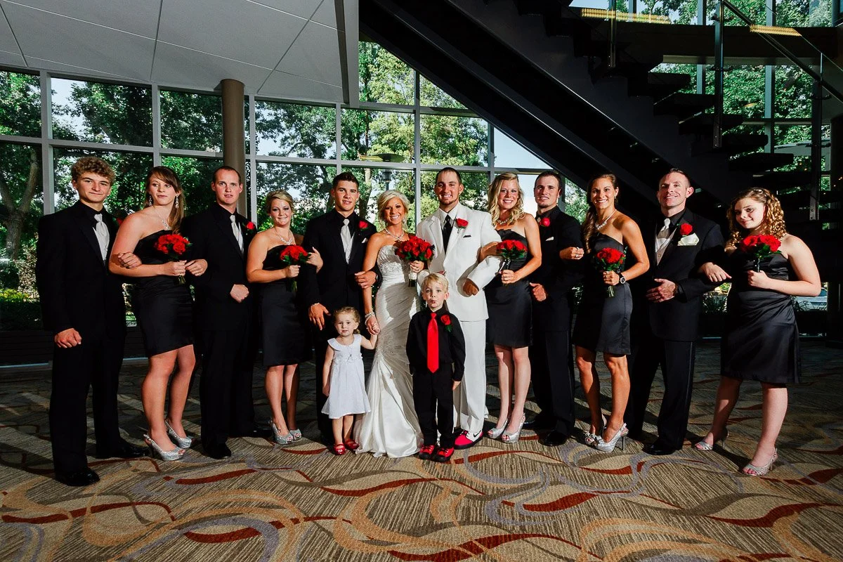 A wedding party poses indoors under a staircase. The bride in white and groom in light suit are centered, surrounded by bridesmaids in black dresses holding red bouquets, groomsmen in black suits, and two children. The mood is joyful.