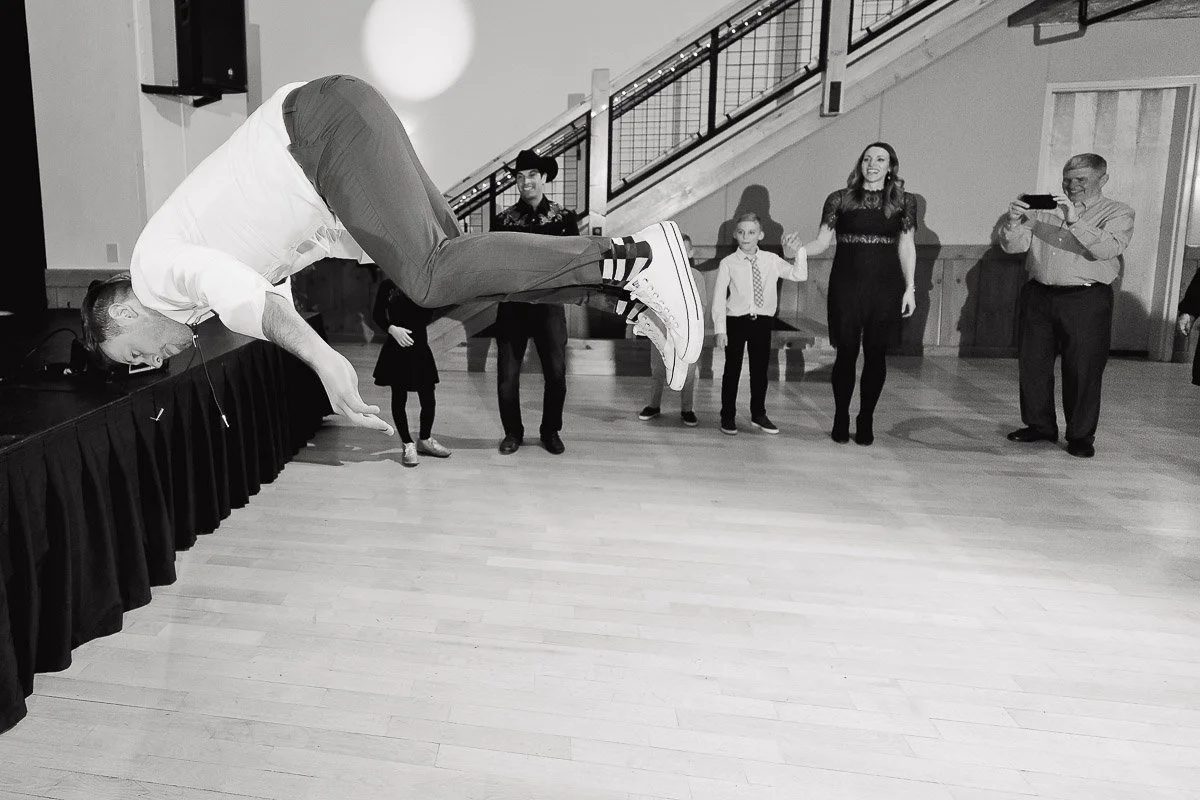 A man in mid-air performing a flip in a room with hardwood floors. Onlookers, including children and adults, watch in amazement. The mood is playful and lively.