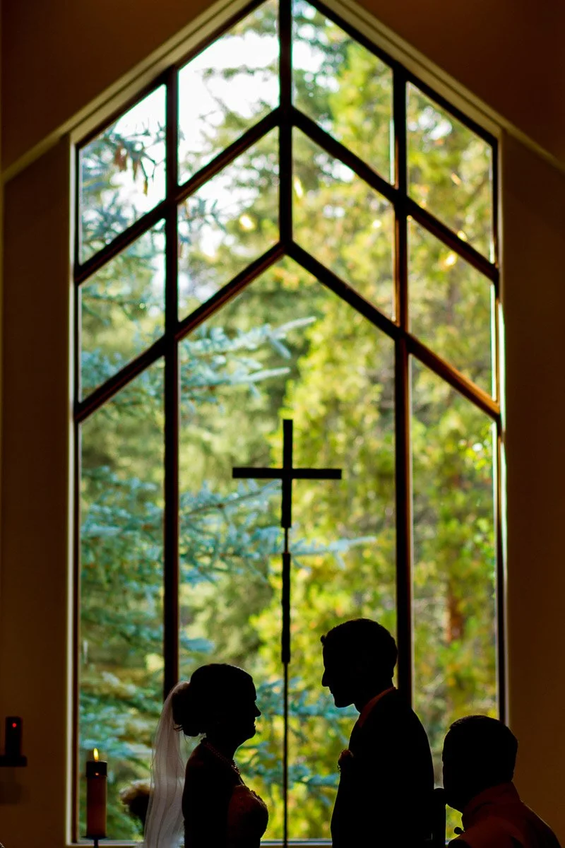 Silhouette of a couple standing in front of a large window with a cross. Sunlight highlights trees outside, creating a serene, spiritual mood during a Vail Interfaith Chapel wedding in Vail Colorado