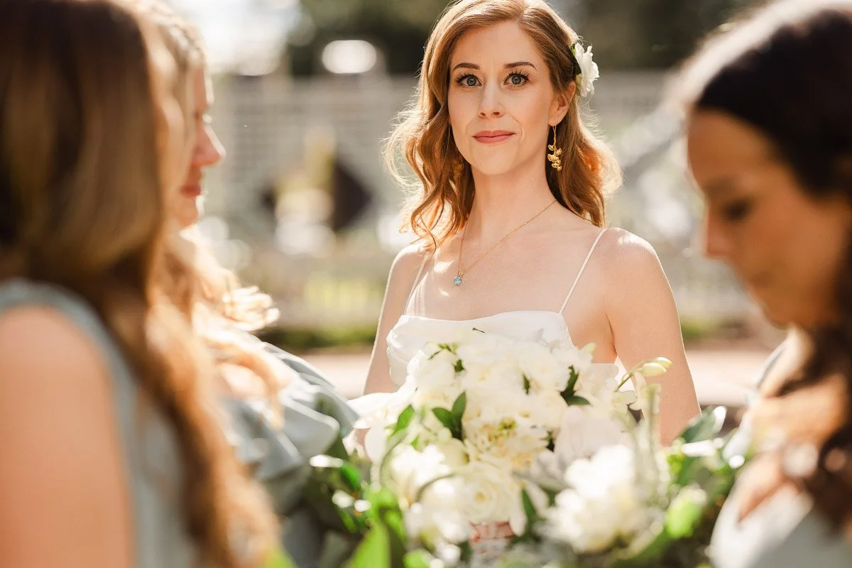 A bride stands in focus, holding white flowers, surrounded by bridesmaids in soft dresses, conveying a serene and joyous wedding atmosphere.