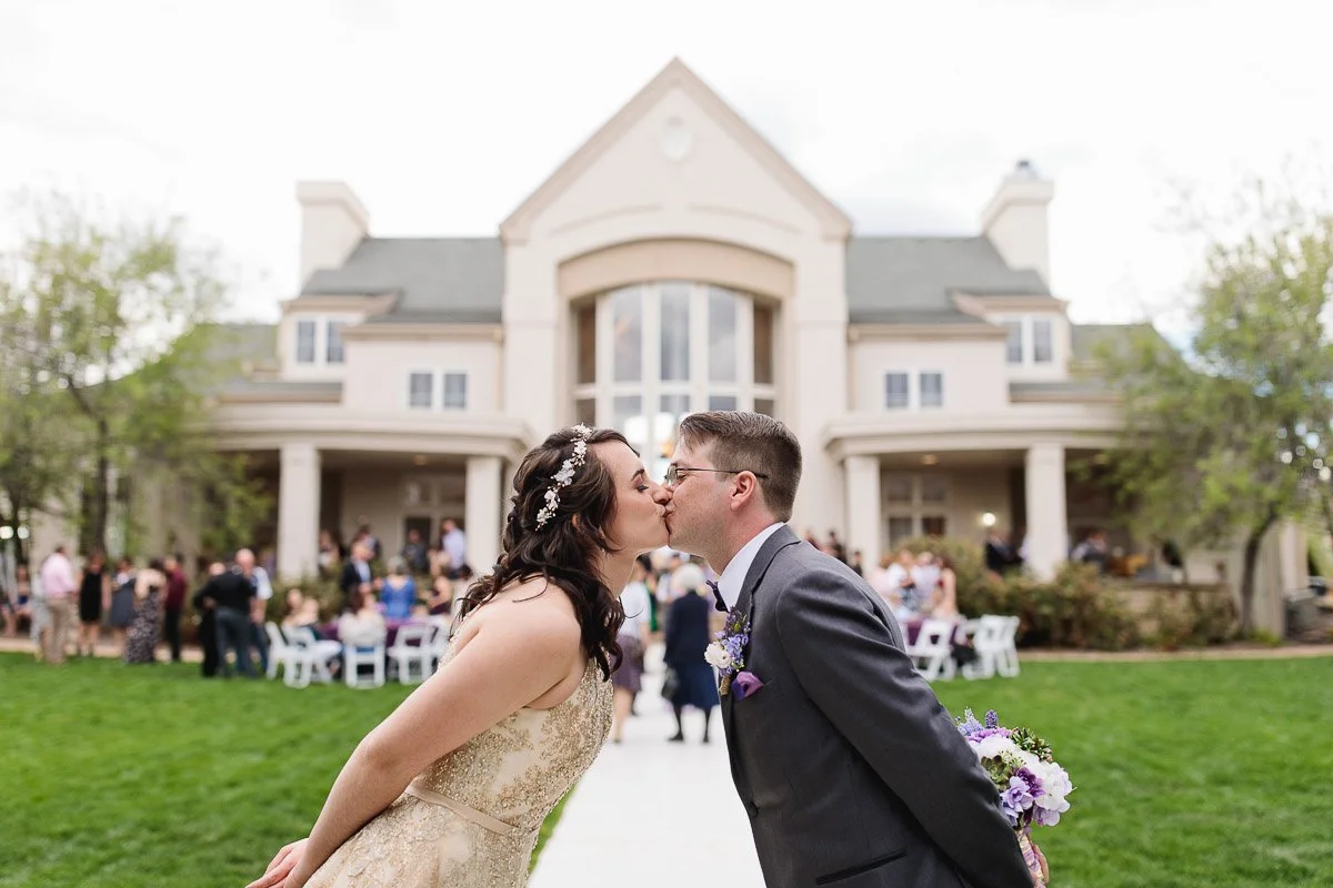 A bride and groom kiss in front of a grand estate. The bride wears a floral headpiece; the groom sports a boutonniere. Guests and greenery surround them.